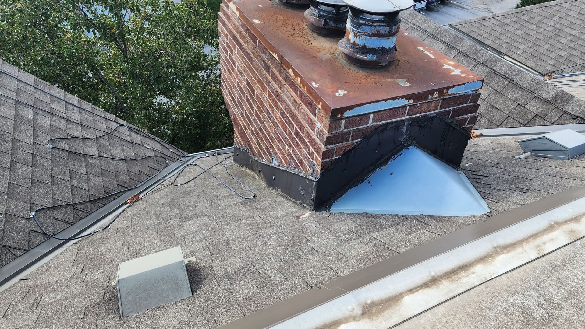 Brick chimney on an Omaha shingle roof with a rusted metal cap and tar smeared over failing base flashing